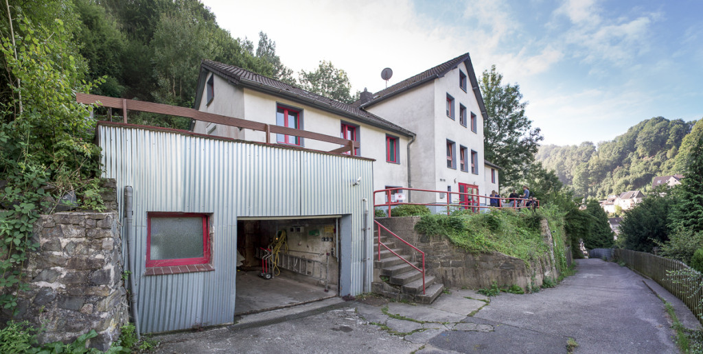 Das Bild zeigt unser Haus in der Totalen vom Wald aus kommend mit der offenen Garage links, dem folgenden Treppenaufgang zum Haus selbst, dem Hang, dem Weg runter zur Hauptstraße mit dem eingezäunten Abhang rechts